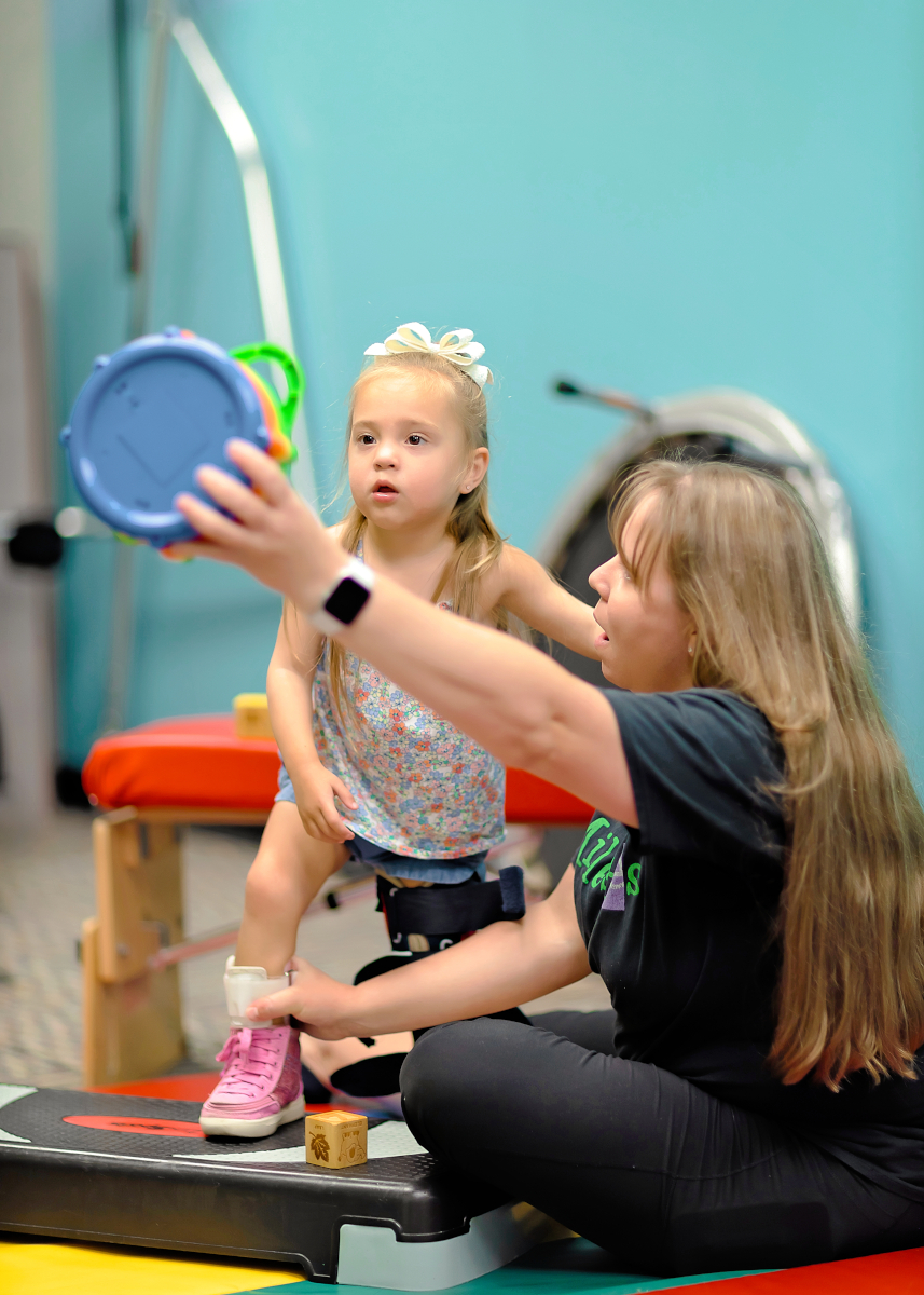 A therapist holds up a colorful music toy with 1 hand and helps support a little girl's leg brace. The little girl focuses on the toy and steps up onto a small platform.