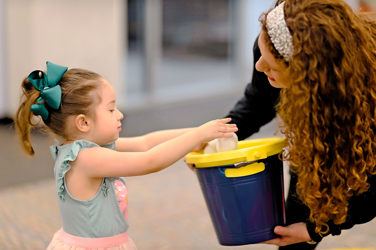 A little girl places balls into a blue bucket that a therapist is holding.
