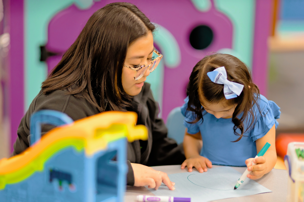 A Therapist watches a little girl draw a circle on a piece of paper with a marker. The little girl is concentrating on the drawing.