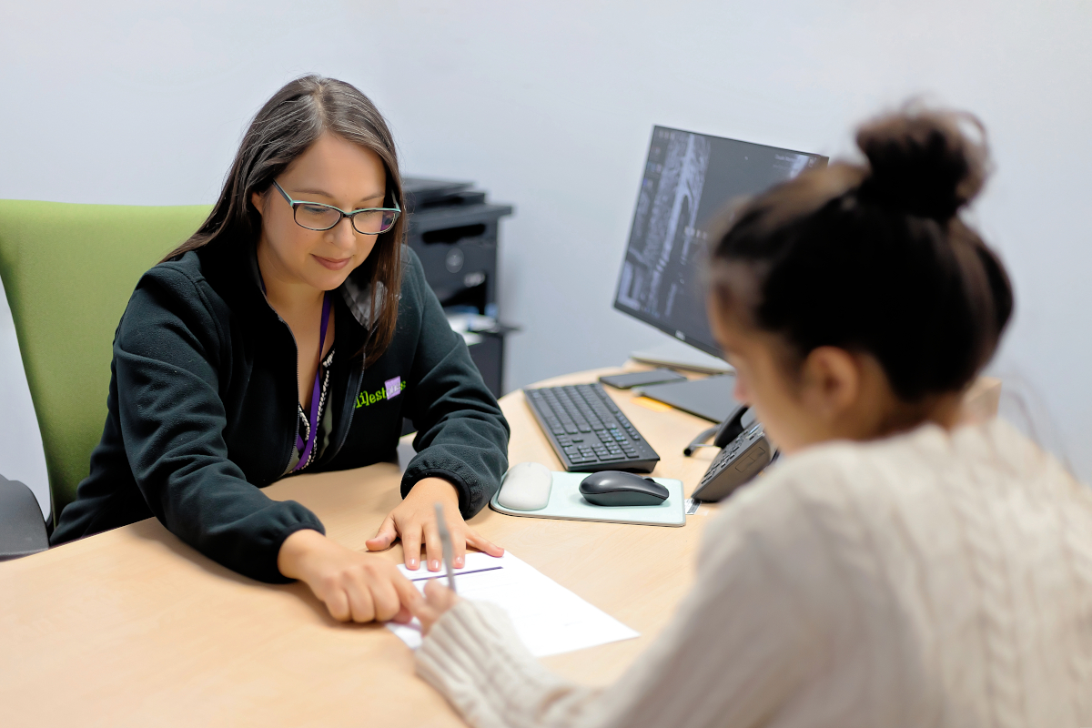 Two women sit at a desk with a piece of paper on the table between them. The woman behind the desk points to the paper while the second woman writes with a pen onto the paper.