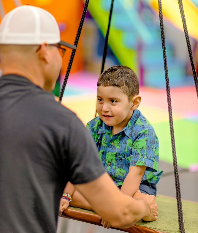 Closer shot of A therapist holds and stabilizes a little boy who is sitting on a modified swing with a flat piece to sit on.