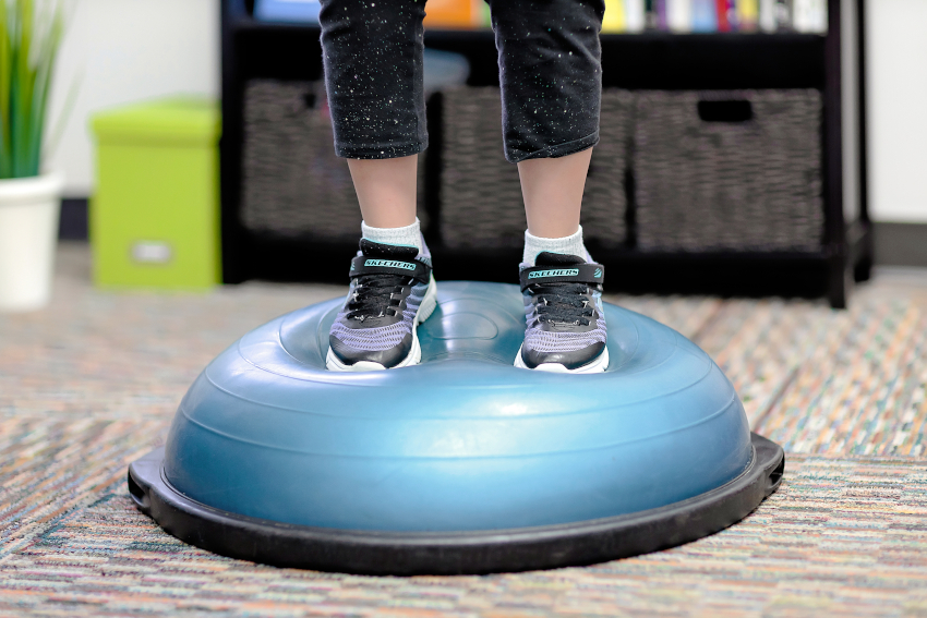 A child wearing sneakers and sparkly pants stands on a squishy exercise ball which is cut in half and placed on a stable base; only the lower legs and feed are visible