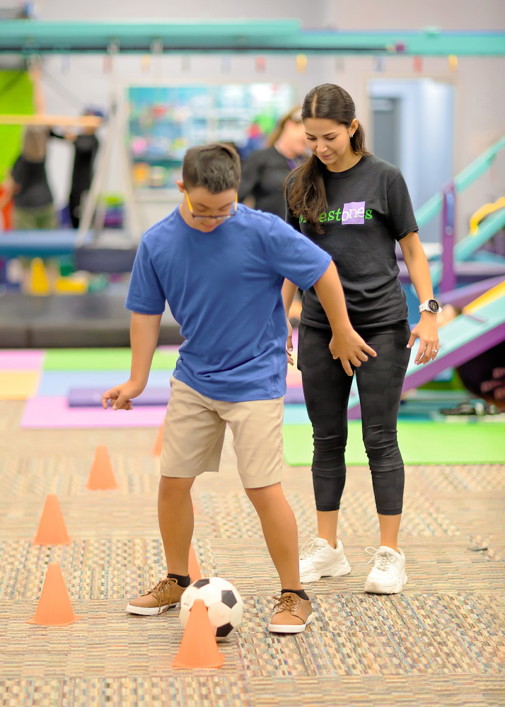 A therapist supervises a boy as he maneuvers a soccer ball between orange cones on the carpeted floor.